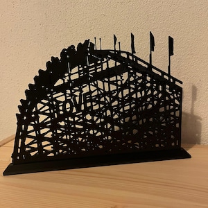 May include: Black metal sculpture of a roller coaster with the word "LOVE" integrated into the design. The coaster features a detailed track and several small flags at the top. The sculpture is displayed on a wooden surface.