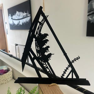 Könnte beinhalten: Eine schwarze Metallskulptur einer Achterbahn, mit einem dreieckigen Rahmen und detailliertem Schienendesign. Die Skulptur zeigt einen Achterbahnwagen mit Passagieren. Das Kunstwerk ist auf einer Holzoberfläche montiert.