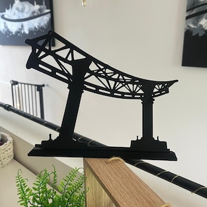 May include: A black metal bridge silhouette, featuring a detailed architectural design. The artwork is displayed on a wooden surface, showcasing the bridge's structure with its support towers and overhead framework. The piece is a decorative item.