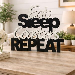 Op de afbeelding: Een zwart-wit houten bord met de woorden "Eat Sleep Coasters Repeat". Het bord staat op een houten tafel. De woorden "Eat" en "Coasters" zijn wit, terwijl "Sleep" en "Repeat" zwart zijn.