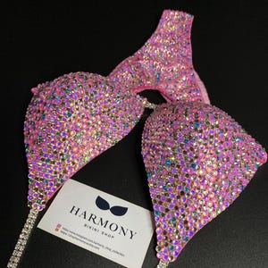 May include: Pink bikini top and bottom with crystal embellishments. The bikini is made with a pink fabric base and covered in iridescent crystals. The bikini top has a halter style and the bottom is a cheeky cut. The bikini is paired with a matching crystal-encrusted strap. The image also includes a business card for "Harmony Bikini Shop" with their website and social media handles.