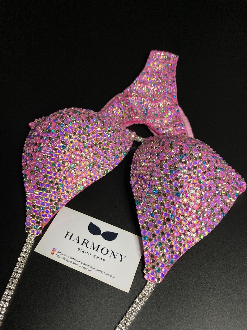May include: Pink bikini top and bottom with crystal embellishments. The bikini is made with a pink fabric base and covered in iridescent crystals. The bikini top has a halter style and the bottom is a cheeky cut. The bikini is paired with a matching crystal-encrusted strap. The image also includes a business card for "Harmony Bikini Shop" with their website and social media handles.