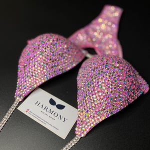 May include: Pink bikini top with crystal embellishments. The top is made of a soft, stretchy fabric and features a halter neck style. The bikini top is decorated with a variety of crystals, including clear, pink, and iridescent stones. The crystals are arranged in a geometric pattern, creating a dazzling effect. The bikini top is perfect for a day at the beach or pool. Harmony Bikini Shop is printed on a card in the image.