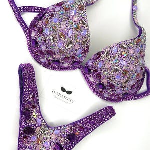 Könnte beinhalten: Ein lila Bikini-Set mit funkelnden Verzierungen. Ober- und Unterteil sind mit einer Vielzahl von schillernden und lila Edelsteinen bedeckt. Der Bikini hat ein klassisches Triangel-Oberteil und einen tief sitzenden Slip. Das Markenetikett lautet "HARMONY BIKINI SHOP".