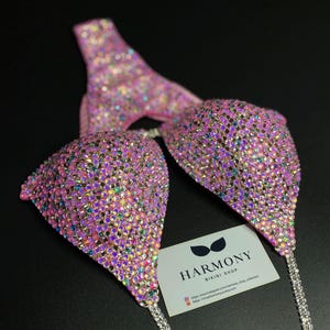 May include: Pink bikini top with iridescent rhinestones and silver chains. The bikini top is from Harmony Bikini Shop. The shop's website and Instagram handle are included on the card.
