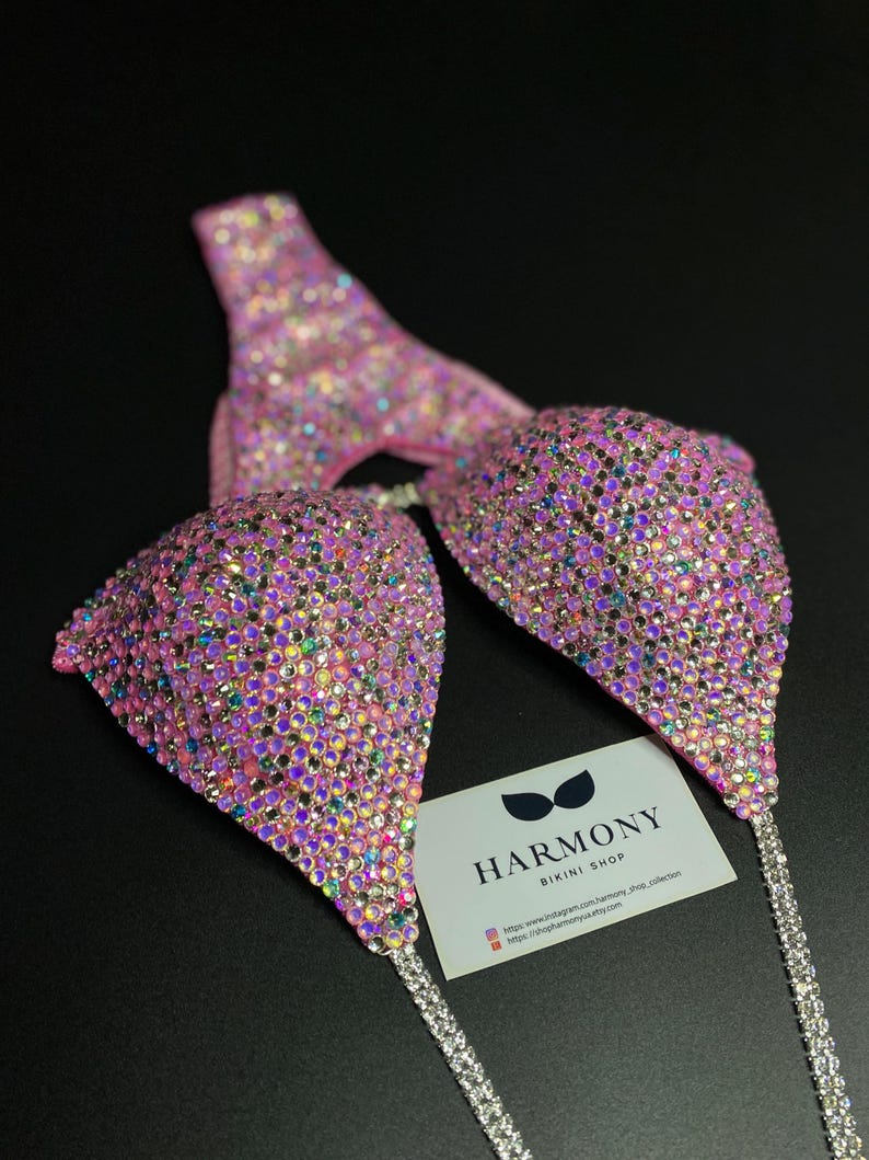 May include: Pink bikini top with iridescent rhinestones and silver chains. The bikini top is from Harmony Bikini Shop. The shop's website and Instagram handle are included on the card.