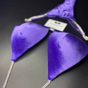Puede incluir: Parte superior de bikini de terciopelo morado con una tira de cadena de cristal. La parte superior tiene un escote pronunciado y una espalda baja.