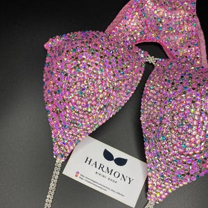 May include: Pink bikini top with iridescent rhinestones and a silver chain. The bikini top is from Harmony Bikini Shop. The text on the card reads "Harmony Bikini Shop" and includes a website address and Instagram handle.