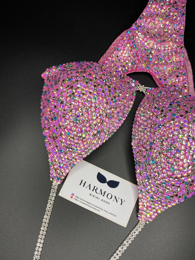 May include: Pink bikini top with iridescent rhinestones and a silver chain. The bikini top is from Harmony Bikini Shop. The text on the card reads "Harmony Bikini Shop" and includes a website address and Instagram handle.
