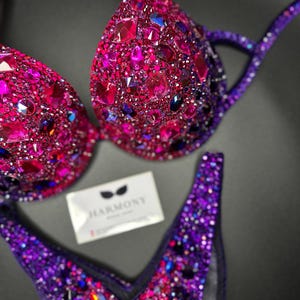 May include: A purple and pink sequined competition bikini with a deep V-neckline. The bikini is decorated with rhinestones and crystals. The brand name "HARMONY" is visible on a white tag.