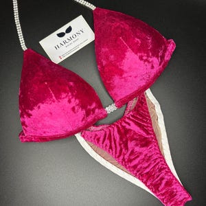 May include: A pink velvet bikini with a crystal-embellished strap and a white mesh lining. The bikini is designed for competition and features a high-cut style.