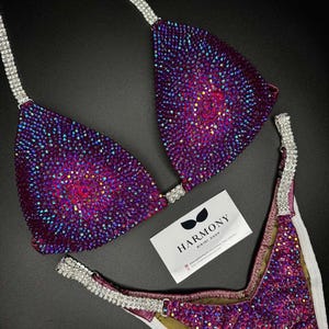May include: Purple and silver sequined bikini competition suit with a white background. The suit has a halter top and a high-cut bottom. The text "HARMONY BIKINI SHOP" is visible on a white card.