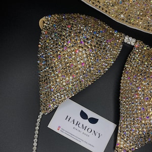 May include: A gold bikini top with crystal embellishments. The top is a triangle style with a thin strap. The bikini top is on a black background with a business card that says "Harmony Bikini Shop" and includes a website and Instagram handle.