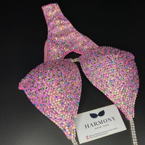 May include: Pink bikini competition suit with crystal embellishments. The suit features a halter top and a bottom with a high cut. The suit is decorated with iridescent crystals. The suit is from Harmony Bikini Shop. The text on the card reads: "Harmony Bikini Shop. https://www.instagram.com/harmony_shop_collection https://shop.harmonybikinis.com"