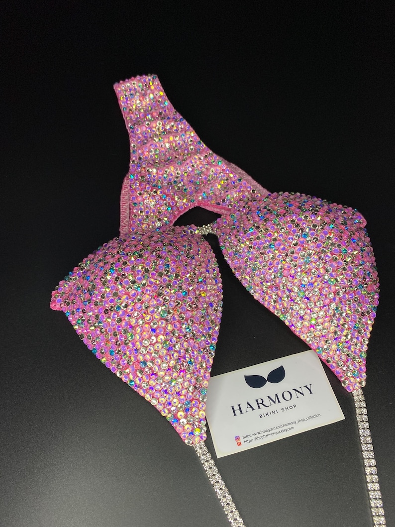May include: Pink bikini competition suit with crystal embellishments. The suit features a halter top and a bottom with a high cut. The suit is decorated with iridescent crystals. The suit is from Harmony Bikini Shop. The text on the card reads: "Harmony Bikini Shop. https://www.instagram.com/harmony_shop_collection https://shop.harmonybikinis.com"