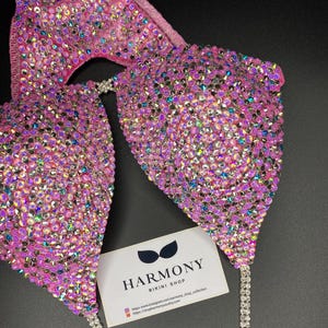 May include: Pink bikini top with iridescent rhinestones and silver chain straps. The top is decorated with a large number of rhinestones. The bikini top is displayed on a black background with a business card that reads "Harmony Bikini Shop".
