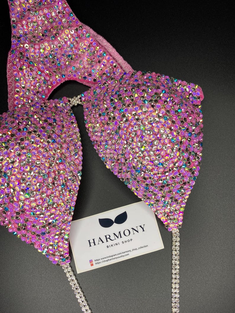 May include: Pink bikini top with iridescent rhinestones and silver chain straps. The top is decorated with a large number of rhinestones. The bikini top is displayed on a black background with a business card that reads "Harmony Bikini Shop".