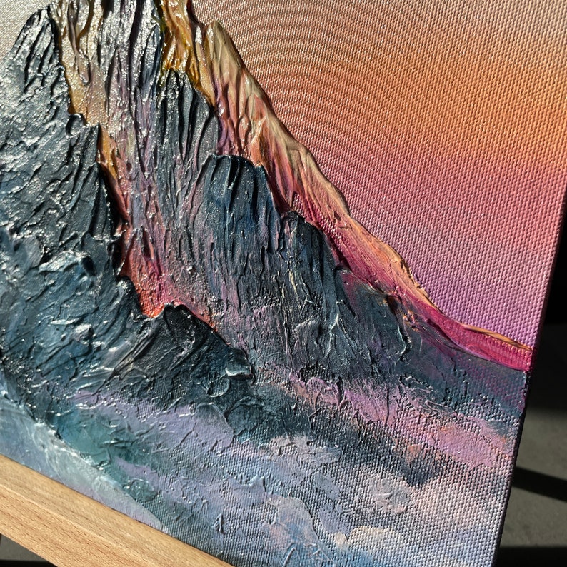 May include: Textured mountain landscape painting with a palette of dark blues, purples, and gold. The artwork features a textured, impasto style, with a gradient sky of pink and orange hues. The canvas is mounted on a wooden easel.