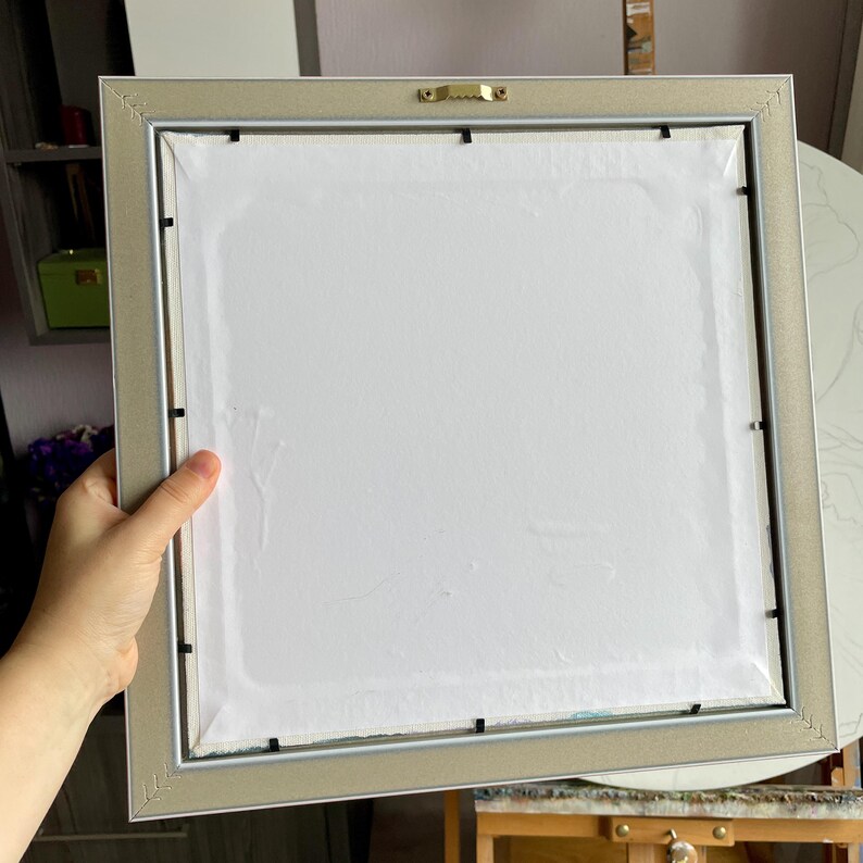 May include: Back of a framed artwork with a silver-colored frame. The artwork is covered with white paper. The frame has a sawtooth hanger for wall mounting. The artwork is being held by a person.