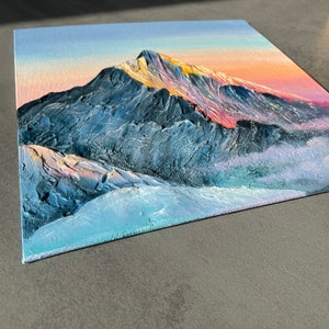 May include: An original oil painting on canvas depicts a mountain range at sunset. The textured artwork features a blue and gray mountain with hints of pink and yellow in the sky. The painting is on a square canvas.