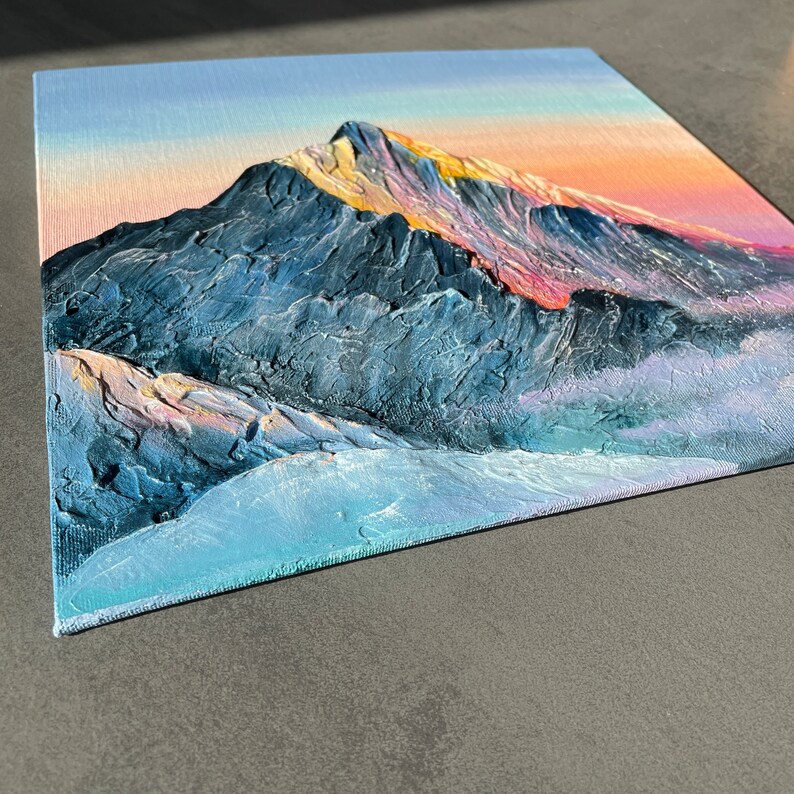 May include: An original oil painting on canvas depicts a mountain range at sunset. The textured artwork features a blue and gray mountain with hints of pink and yellow in the sky. The painting is on a square canvas.