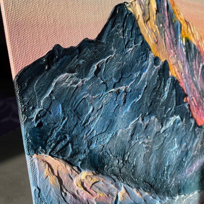 May include: Textured oil painting of a mountain range. The artwork features a dark blue mountain with a textured surface, set against a pink and orange sky. The canvas is stretched and mounted, showcasing the side profile of the artwork.