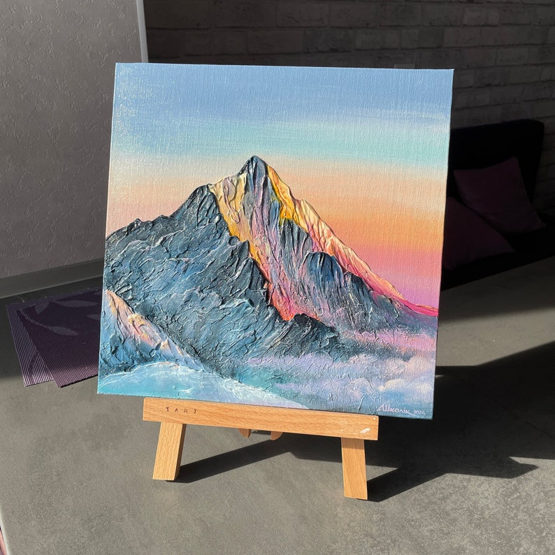May include: Square canvas art of a mountain range with a textured, impasto style. The mountain is painted in shades of blue and gray, with the peak highlighted in yellow and pink. The sky transitions from blue to orange. The artwork is displayed on a small wooden easel.