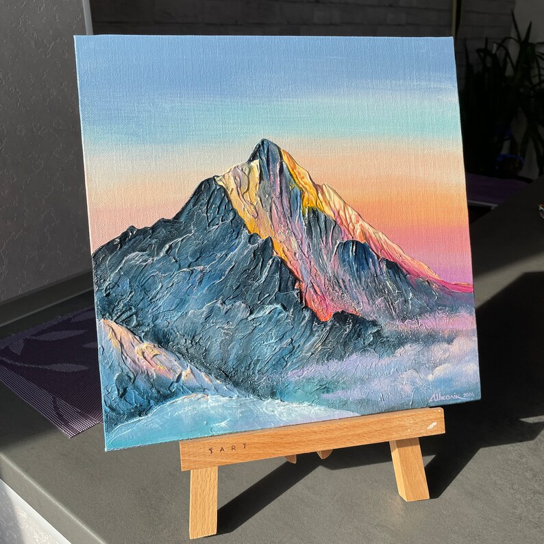 May include: An original oil painting on canvas depicts a mountain range at sunset. The textured artwork features a blue and gray mountain with golden peaks, set against a sky of pink, orange, and blue hues. The painting is displayed on a small wooden easel.