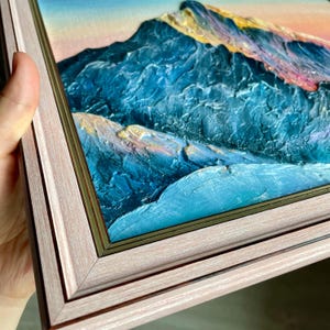 May include: A framed oil painting of a mountain range. The artwork features textured brushstrokes in shades of blue, teal, and gold, with a pink and yellow sky. The frame is a light pink color with a gold inner edge.