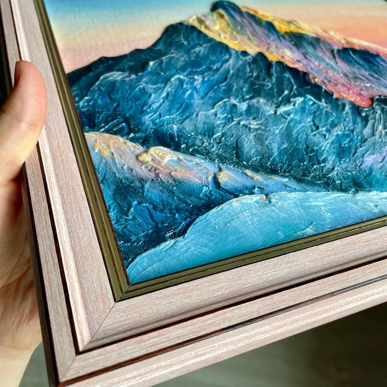 May include: A framed oil painting of a mountain range. The artwork features textured brushstrokes in shades of blue, teal, and gold, with a pink and yellow sky. The frame is a light pink color with a gold inner edge.