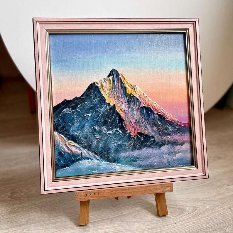 May include: An oil painting of a mountain peak with a pink and gold frame. The mountain is painted with shades of blue, purple, and gold, with a pink and orange sky in the background. The painting is displayed on a small wooden easel.