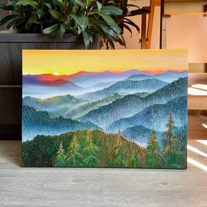 以下が含まれることがあります： 夕焼けの山岳風景を描いた油絵。空は黄色からオレンジ色に変わり、遠くの山々の上に太陽が顔をのぞかせています。前景には緑の木々があり、背景には青と紫の山々が重なっています。