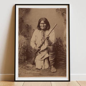 Op de afbeelding: Sepia-getinte ingelijste print met een portret van een knielende Indiaanse man die een geweer vasthoudt. Hij draagt een lichtgekleurde tuniek en kniehoge laarzen. De achtergrond bevat woestijnvegetatie, wat een historisch en cultureel thema creëert.