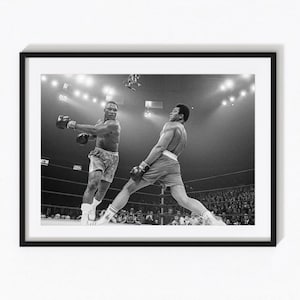 May include: A black and white framed photograph depicting two boxers in a ring during a match. One boxer is throwing a punch while the other leans back to dodge. The scene is captured under bright arena lights with a crowd visible in the background.