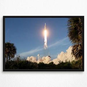 以下が含まれることがあります： 白いロケットが澄んだ青空に向かって垂直に上昇し、炎と排気ガスの明るい軌跡を残しています。発射場からは厚い白い煙の雲が立ち上り、両側にあるヤシの木の濃い緑色のシルエットがその光景を縁取っています。