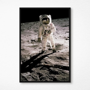 Pode incluir: Uma impressão emoldurada de um astronauta na lua. O astronauta está usando um traje espacial branco com um patch da bandeira americana. A superfície lunar é cinza e empoeirada, com a sombra do astronauta. A impressão está em uma moldura preta.