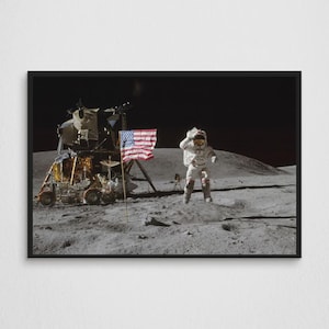 Op de afbeelding: Een ingelijste print van de iconische afbeelding van een astronaut op de maan, met de Amerikaanse vlag en de maanlander op de achtergrond. De astronaut draagt een wit ruimtepak en springt op het maanoppervlak. De vlag is rood, wit en blauw.