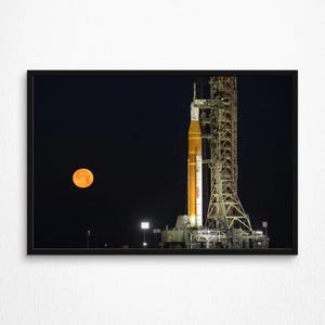 Könnte beinhalten: Gerahmter Druck einer Rakete auf einer Startrampe bei Nacht, mit einem großen orangefarbenen Mond im Hintergrund. Die Rakete ist orange und weiß, mit dem NASA-Logo sichtbar. Der Startturm ist eine komplexe Metallkonstruktion.