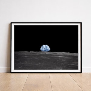 May include: Framed print featuring the iconic "Earthrise" photograph. The image shows the Earth, a blue and white sphere, rising above the lunar surface. The black frame surrounds the image, which is set against a black background, with a wooden floor below.