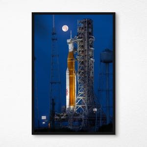 以下が含まれることがあります： 夜の打ち上げ台にあるNASAのロケットの額入りプリント。満月が空に見えます。ロケットは白とオレンジ色で、発射塔は複雑な金属構造です。背景は濃い青色です。