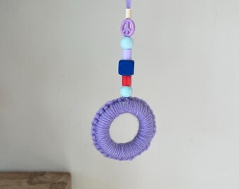 May include: A handmade lavender-colored crochet ring ornament with a string of colorful beads. The beads include a peace sign, blue, red, and purple accents. The ornament is suspended from a matching lavender string.