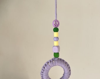 May include: A lavender macrame ring ornament with a matching cord for hanging. The ornament is adorned with a string of beads in green, yellow, and purple, including a peace sign charm. A decorative item for a home or car.