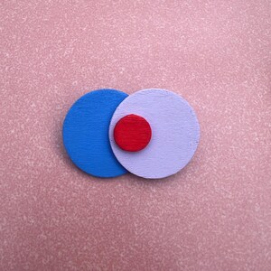Könnte beinhalten: Ein abstraktes Design mit drei sich überlappenden Kreisen. Ein großer blauer Kreis überlappt einen lavendelfarbenen Kreis, mit einem kleineren roten Kreis in der Mitte. Der Hintergrund ist eine strukturierte rosa Oberfläche.