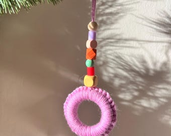 May include: A handmade pink macrame ring ornament with a string of colorful wooden beads. The beads include light purple, pink, orange, green, and red. The ornament is hanging from a pink string, with a green leafy branch in the background.