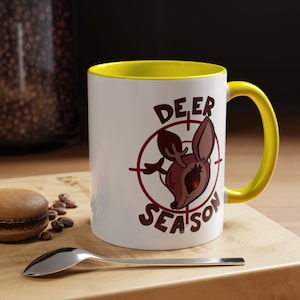 Puede incluir: Taza de cerámica blanca con interior y asa amarillos. La taza presenta una cabeza de ciervo de dibujos animados dentro de una diana, con las palabras "DEER SEASON" en letras marrones. Una cuchara y un macaron están sobre una superficie de madera.
