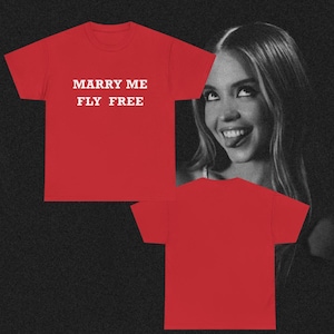 以下が含まれることがあります： 「MARRY ME FLY FREE」の白い文字が入った赤いTシャツ。画像には、背景に女性の白黒写真も含まれています。