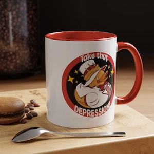 Puede incluir: Taza de cerámica blanca con interior y asa rojos. La taza presenta una imagen de dibujos animados de un pato chef con el texto "Take that DEPRESSION!" en un diseño circular rojo y negro. La taza está sobre una superficie de madera.