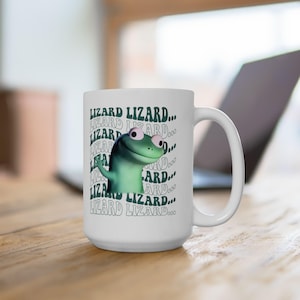 May include: White ceramic mug featuring a cartoon green lizard with large eyes and the text "LIZARD LIZARD..." repeated around the lizard. The mug has a large handle and is set against a blurred background.
