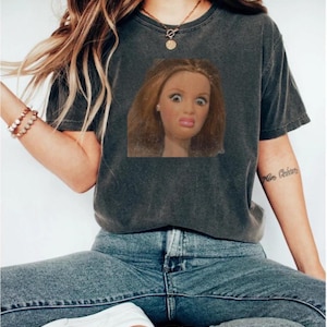 May include: Dark grey t-shirt featuring a doll's face with a surprised expression. The doll has light brown hair and is making a funny face. The shirt is paired with blue jeans and white shoes.