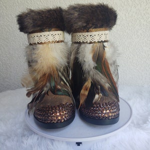 May include: A pair of brown suede boots with faux fur trim, embellished with feathers and rhinestones. The boots have a lace trim and a gold accent.